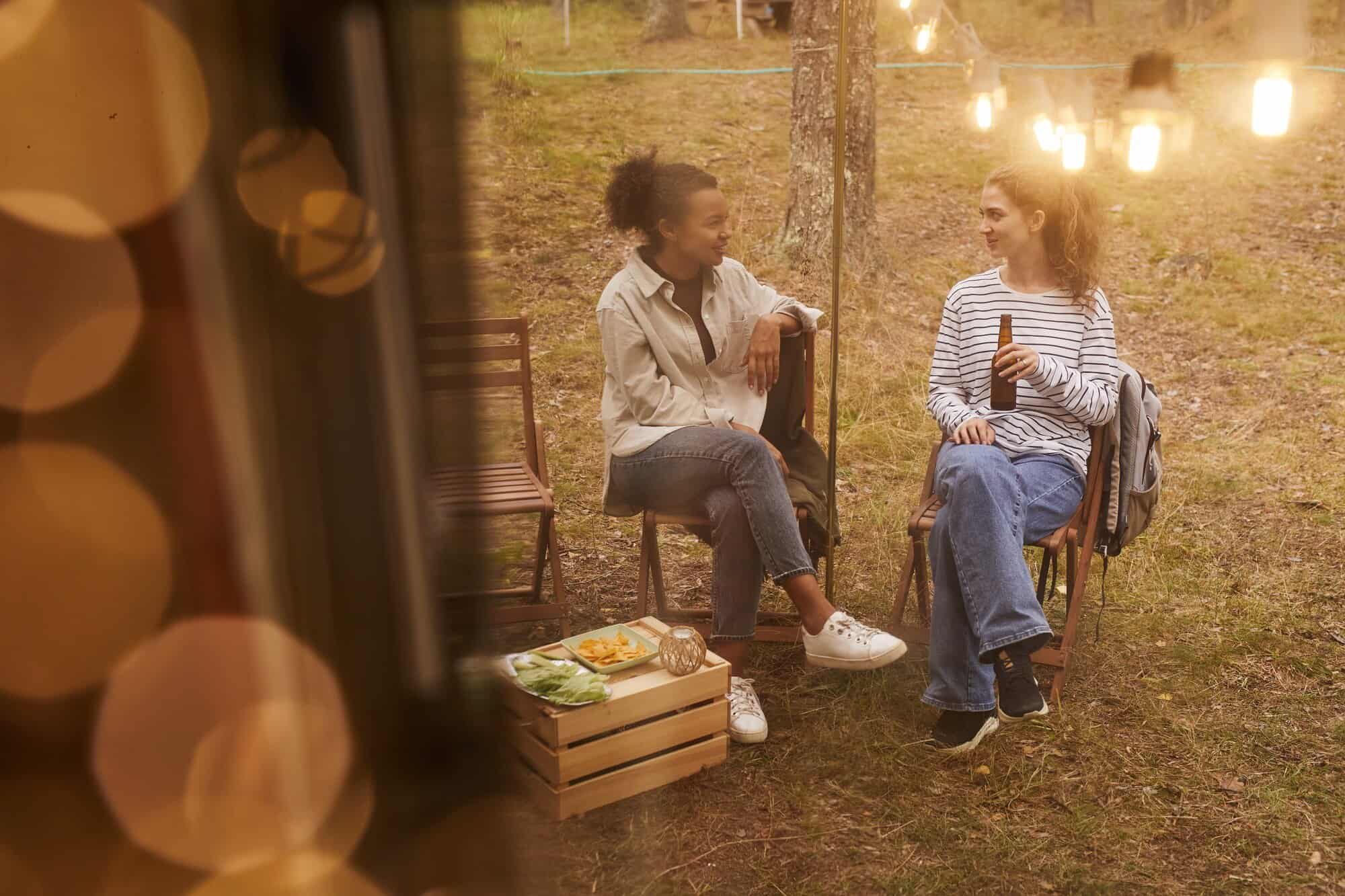 Relaxed women chatting outdoors at a sunset gathering, enjoying food and drinks, surrounded by string lights and nature.